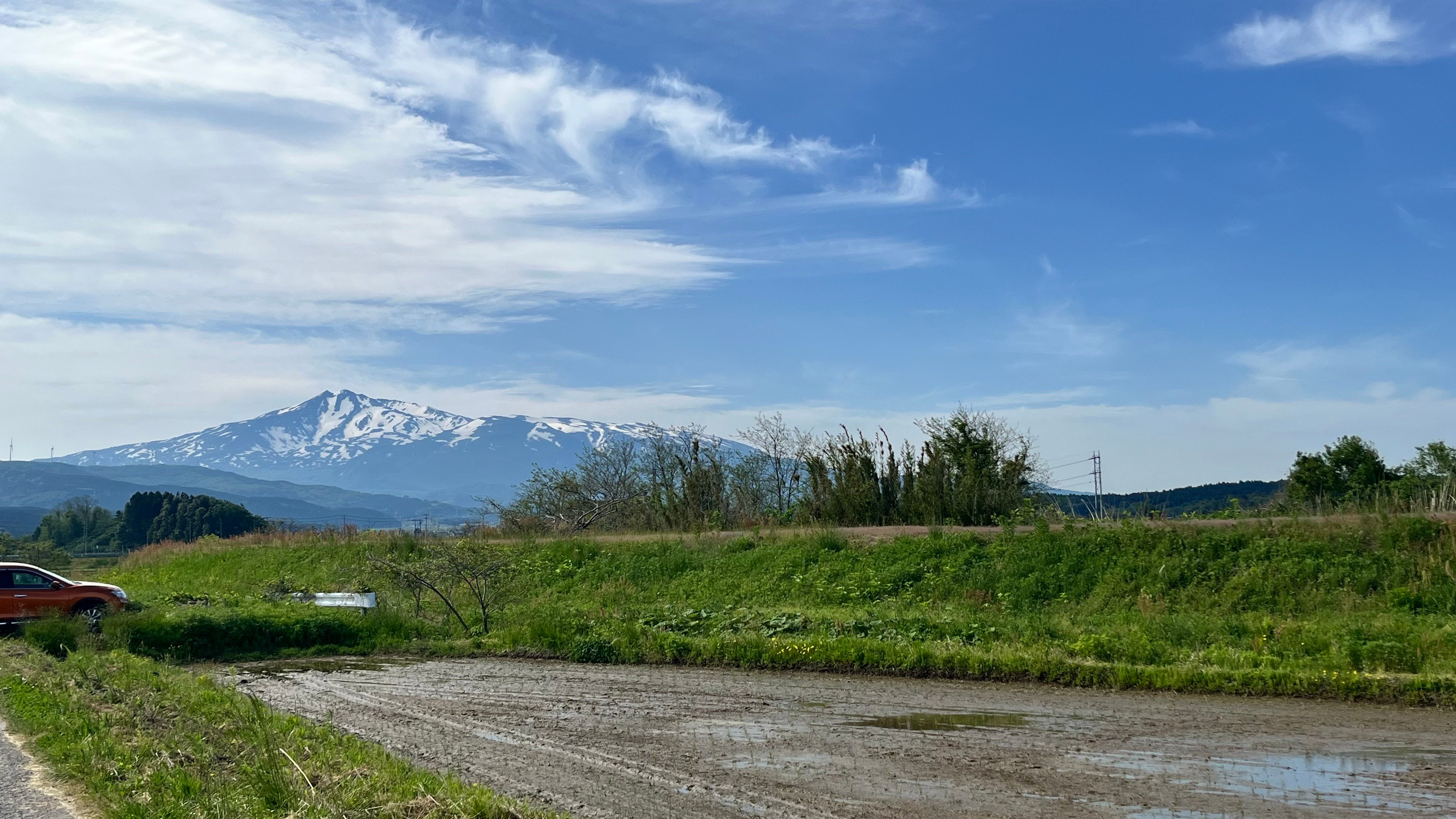 Rural rice farming landscape in Akita Japan with snow-capped Mount Chokai in background, muddy farm track and spring paddy fields