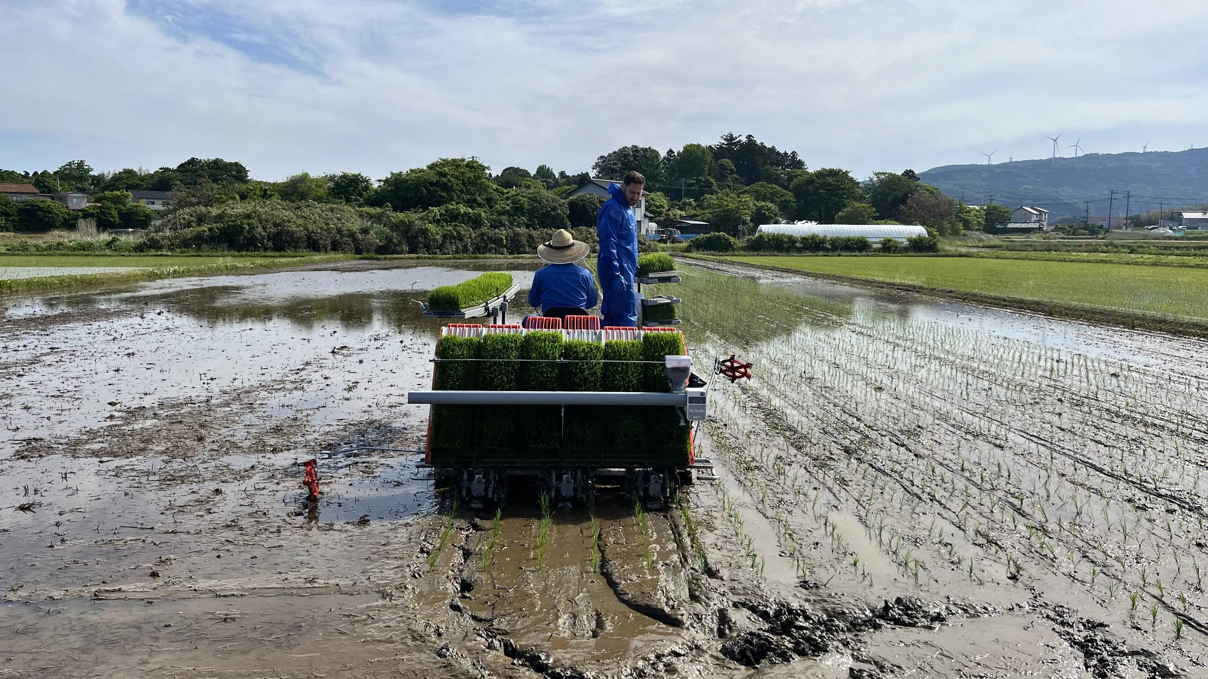 Greenda CEO Chadi Nemr riding a rice transplanter through flooded paddies in Nikaho, Akita, Japan, alongside a local farmer during the spring 2026 planting season — seedling mats loaded and rows of young rice plants visible in the mud behind the machine.
