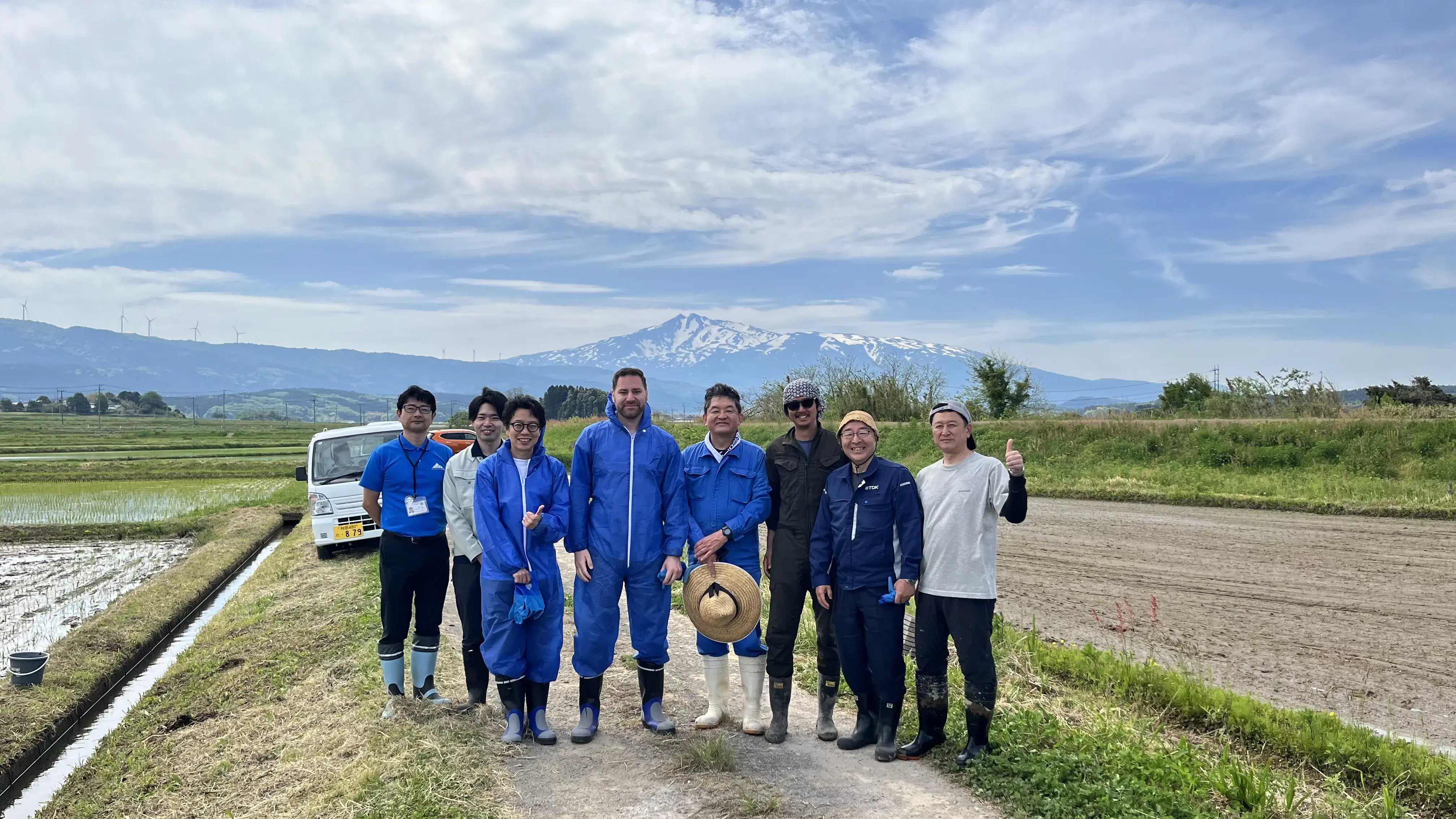 International agricultural research team visiting Japanese rice farm with flooded paddies and snow-capped mountain backdrop in rural Japan
